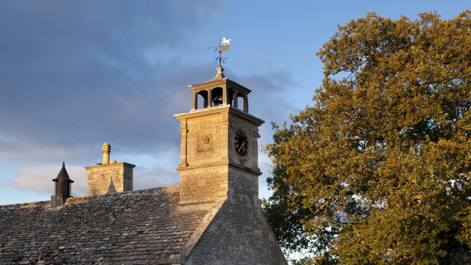 Warm evening light on a close-up of a clock tower on top of a village hall and a tree displaying signs of autumn in its orangey leaves